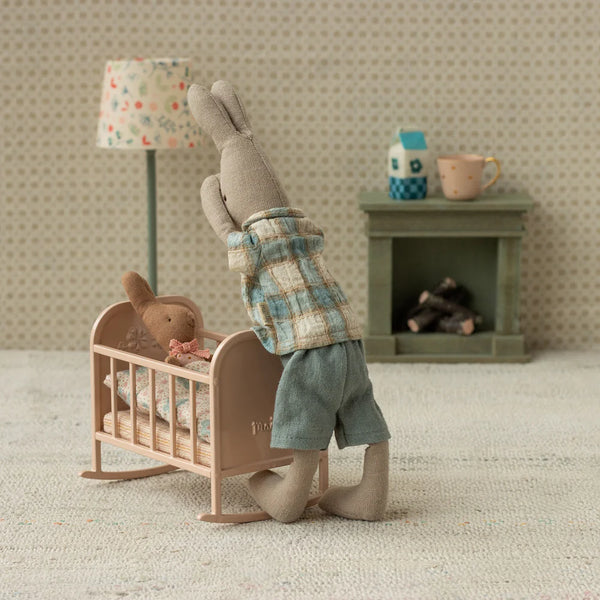 Small rabbit-like creature in a plaid shirt and shorts standing next to a baby crib with a teddy bear inside, in a room with a lamp and fireplace.