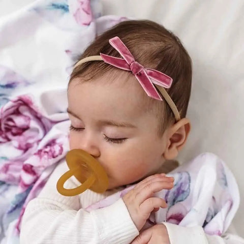 Baby with a pacifier and pink headband lying on a floral blanket