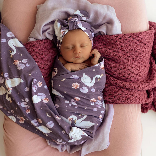 Newborn baby wrapped in a purple swaddle with swan pattern, wearing a matching headband, lying on a pink blanket.