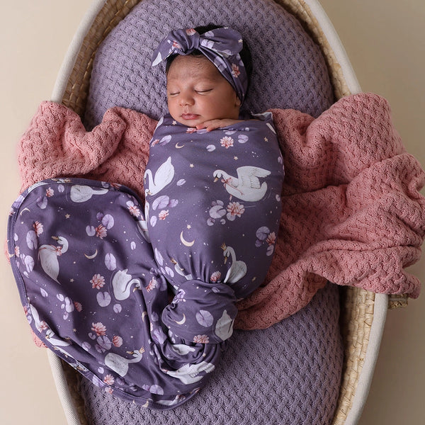 Newborn baby wrapped in a purple blanket with animal patterns, lying on a pink blanket in a basket.