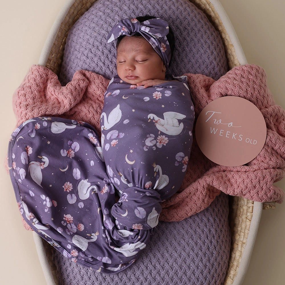 Newborn baby wrapped in a purple blanket with swan pattern, wearing a matching headband, in a crib with pink and purple blankets.