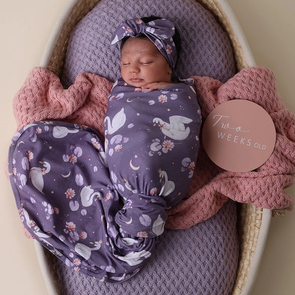 Newborn baby wrapped in a purple blanket with swan pattern, wearing a matching headband, in a crib with pink and purple blankets.