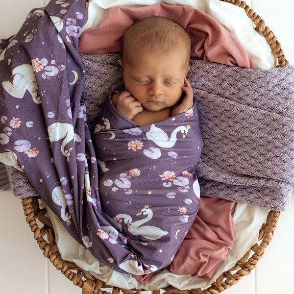 Newborn baby swaddled in a purple blanket with swan design in a wicker basket.
