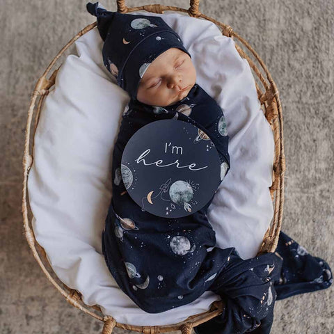 Newborn baby wrapped in a navy blue swaddle with celestial patterns, lying on a white blanket in a wicker basket.