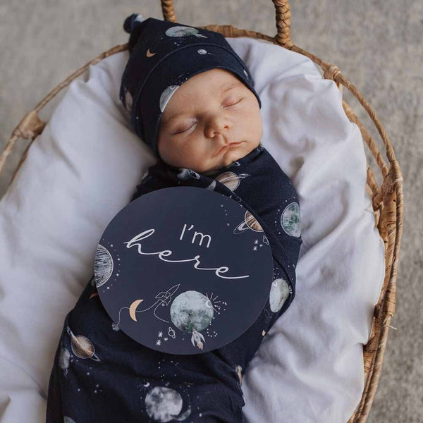 Newborn baby wrapped in a navy blanket with space-themed design, wearing a matching hat, and lying on white fabric in a wicker basket.