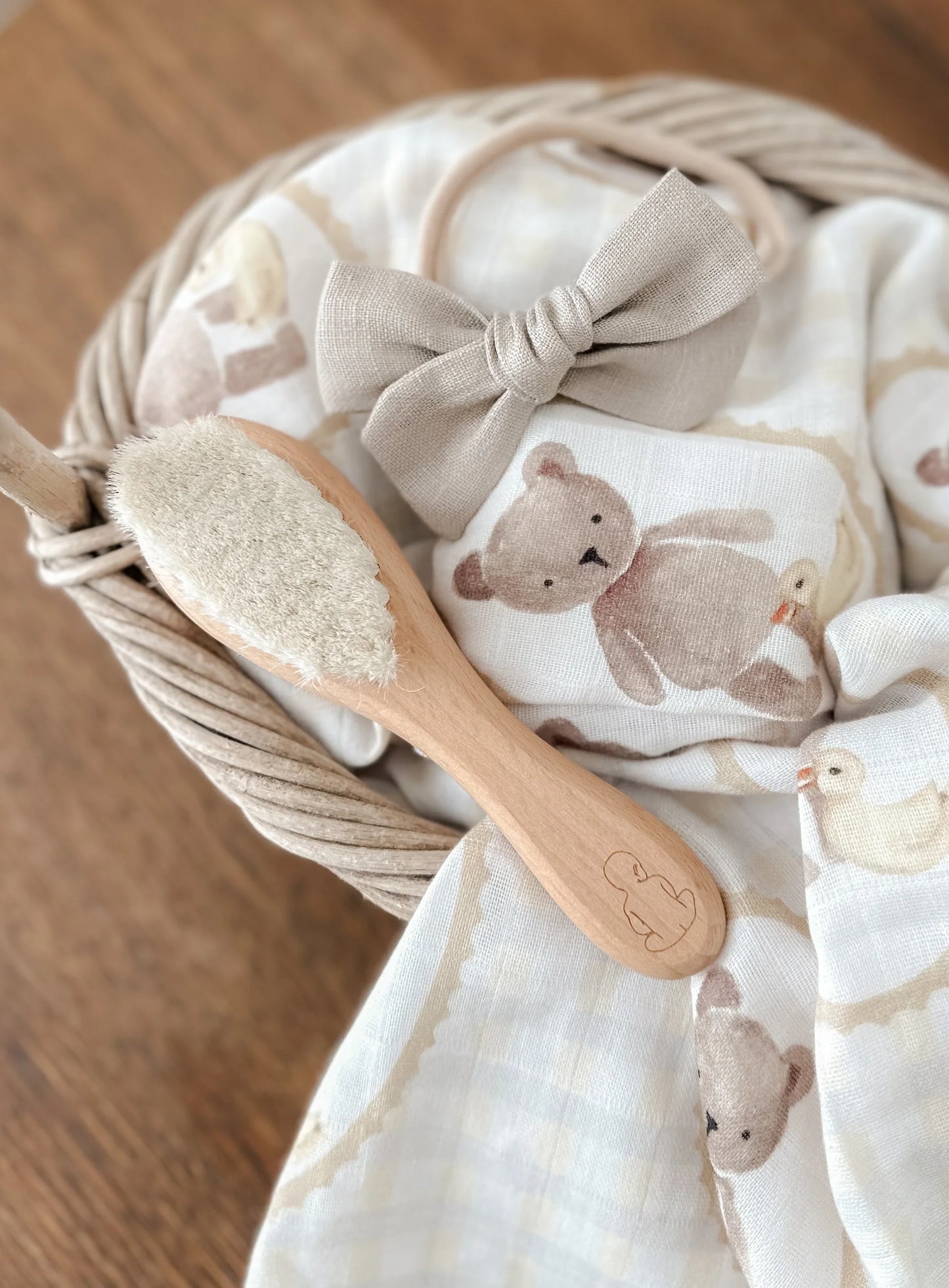 Baby blanket with teddy bear pattern, wooden brush, and bow in a basket on a wooden surface