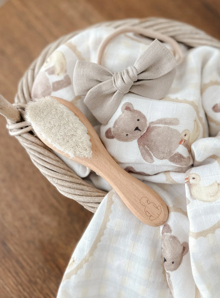 Baby blanket with teddy bear pattern, wooden brush, and bow in a basket on a wooden surface