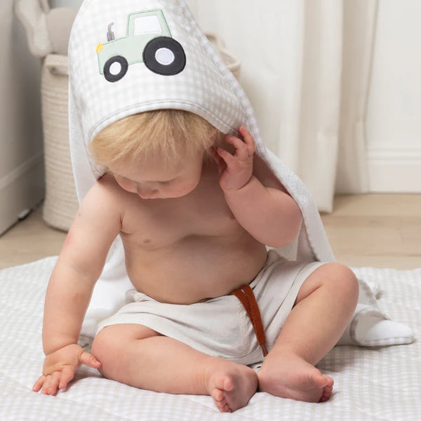 Baby sitting on a bed wearing a hooded towel with a tractor design.