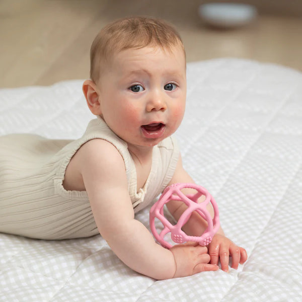 Baby holding a pink teething ball on a white blanket