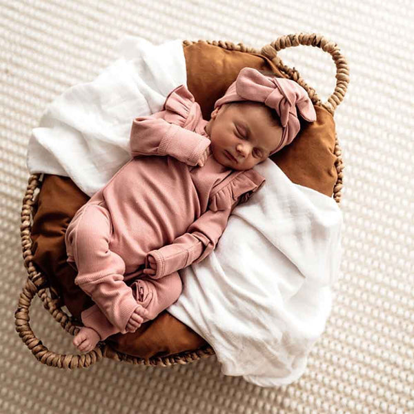 Newborn baby in a pink outfit and headband sleeping in a brown basket with white blanket.