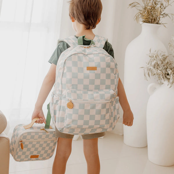 Child wearing a checkered backpack and suitcase in a room with white walls and decor.