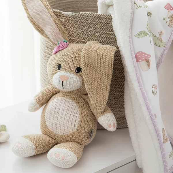 Knitted bunny toy with a pink bow next to a textured basket and floral-patterned blanket.