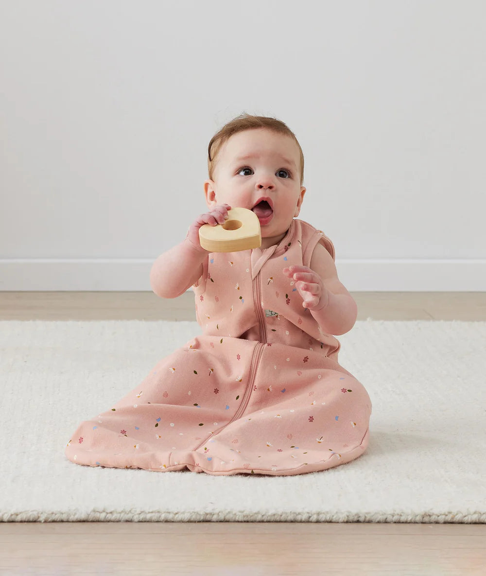 Baby in a pink outfit holding a wooden heart toy on a light-colored floor.