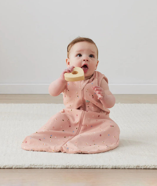 Baby in a pink outfit holding a wooden heart toy on a light-colored floor.