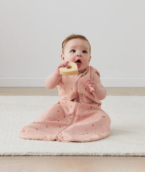 Baby in a pink outfit holding a wooden heart toy on a light-colored floor.