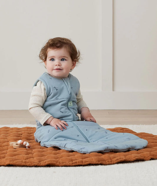 Baby sitting on a wooden floor wearing a blue sleeping bag with a white wall in the background
