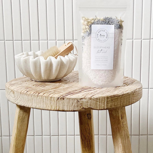 Wooden stool with a white bowl and a bag of bath salts against a tiled wall.