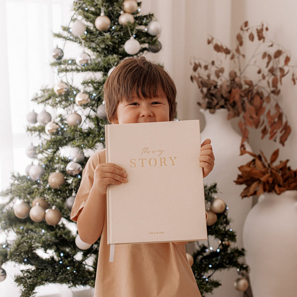 Child holding a book titled 'My Story' in front of a decorated Christmas tree.