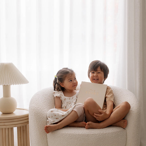 Two children sitting on a chair holding a book in a softly lit room.