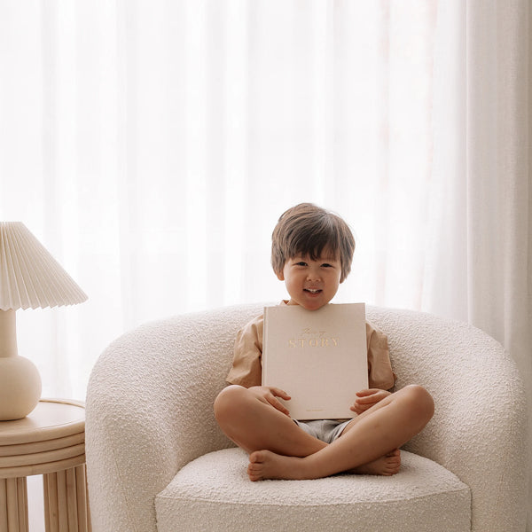 Child sitting on a white armchair holding a book with a white curtain in the background