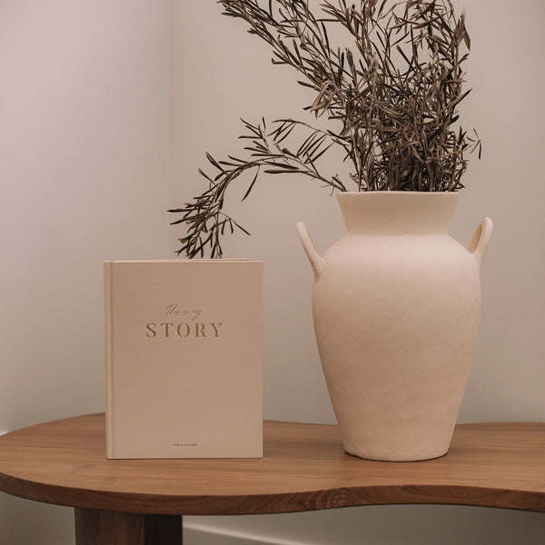 Beige vase with dried branches and a beige book titled 'This is my STORY' on a wooden surface.