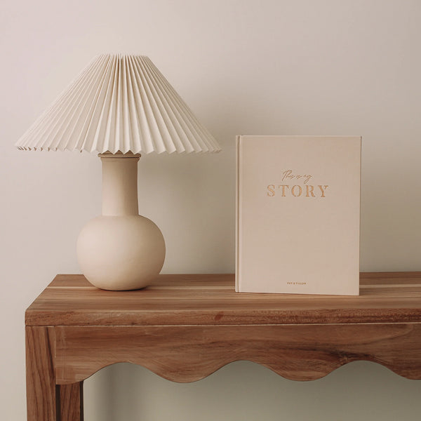 Beige lamp and book titled 'My Story' on a wooden table against a plain wall.