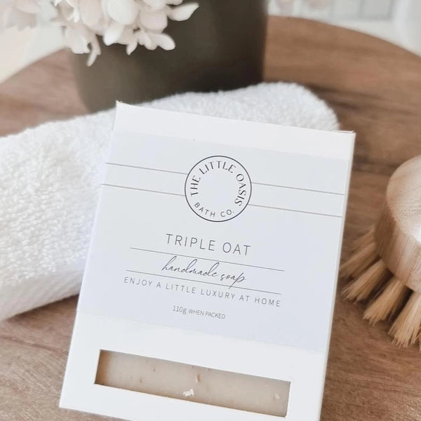 Triple Oat soap packaging on a wooden surface with a towel and brush in the background