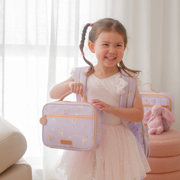 Young girl holding a purple lunch bag with pink accents in a bright room.