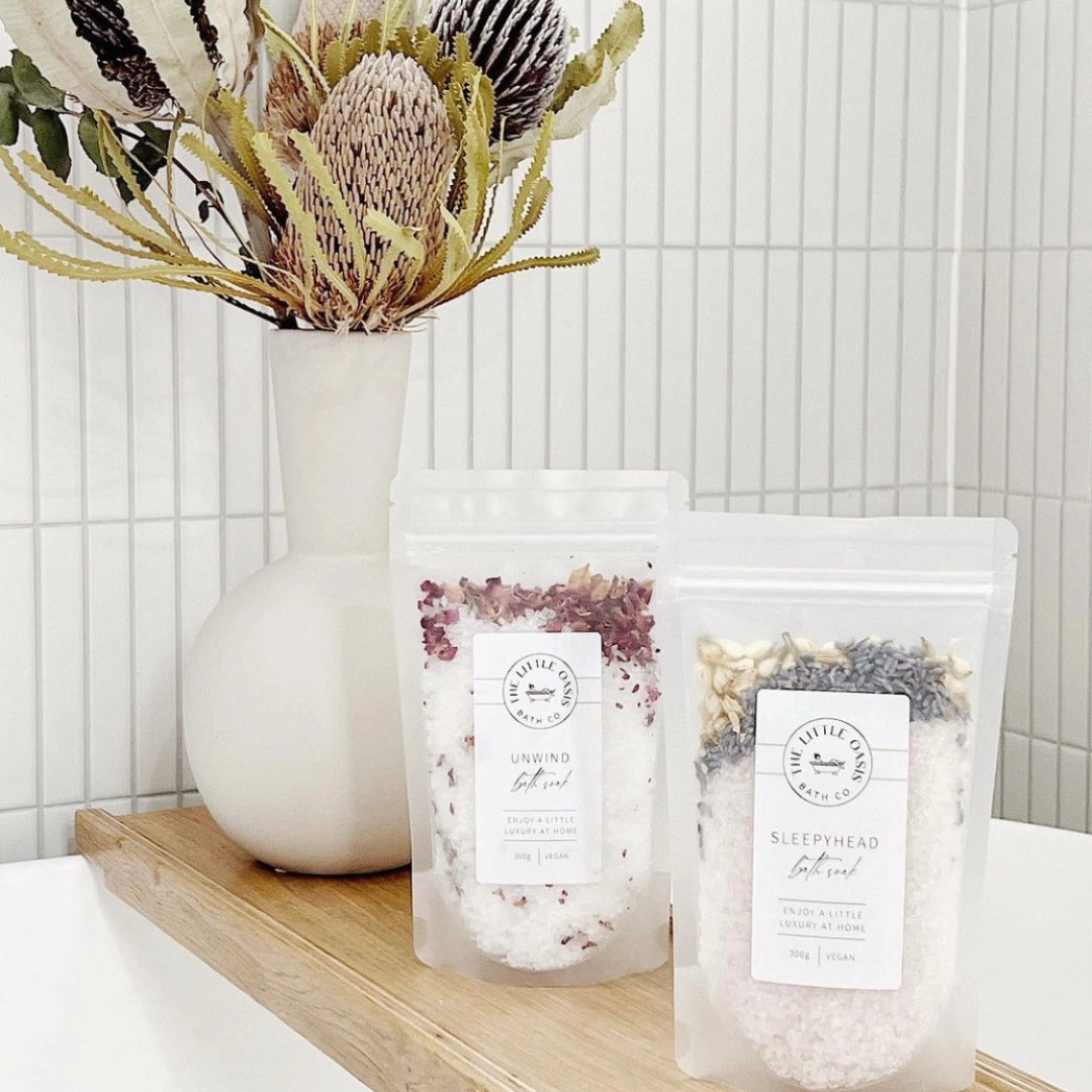 Two packages of bath salts on a wooden shelf with a vase of flowers in the background.