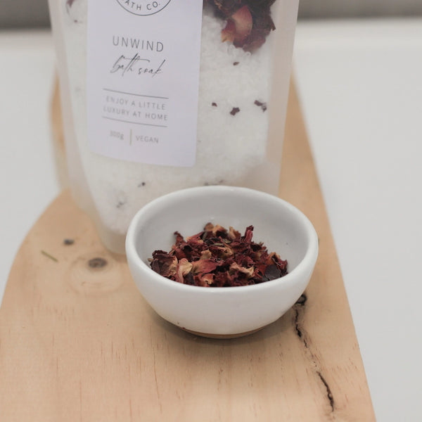 Bath salt package labeled 'Unwind' with a small bowl of dried herbs on a wooden board.
