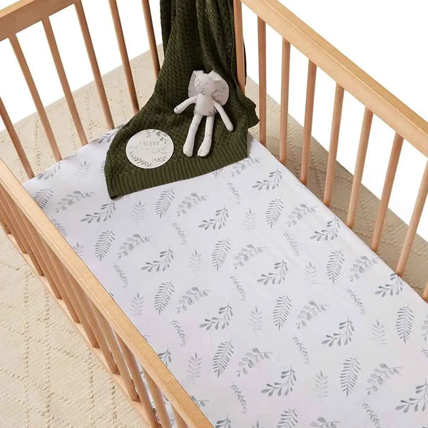 Crib with a green knitted blanket and gray elephant toy on a white sheet with leaf pattern.