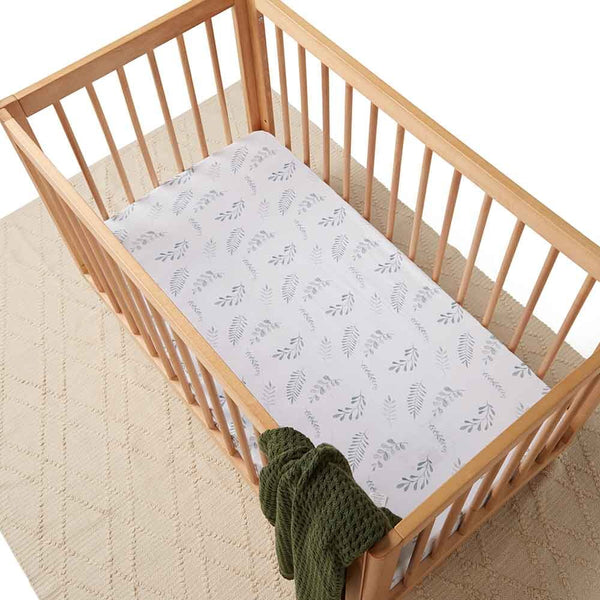 Wooden crib with a white mattress featuring a leaf pattern and a green blanket on a beige carpet.