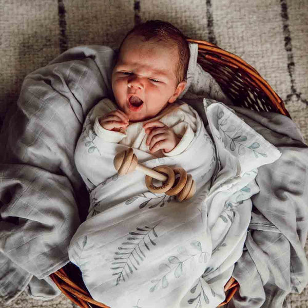 Newborn baby wrapped in a swaddle with a wooden teething ring, lying on a textured surface.