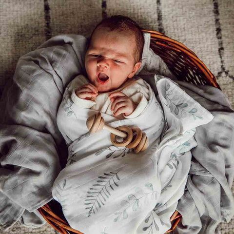 Newborn baby wrapped in a swaddle with a wooden teething ring, lying on a textured surface.