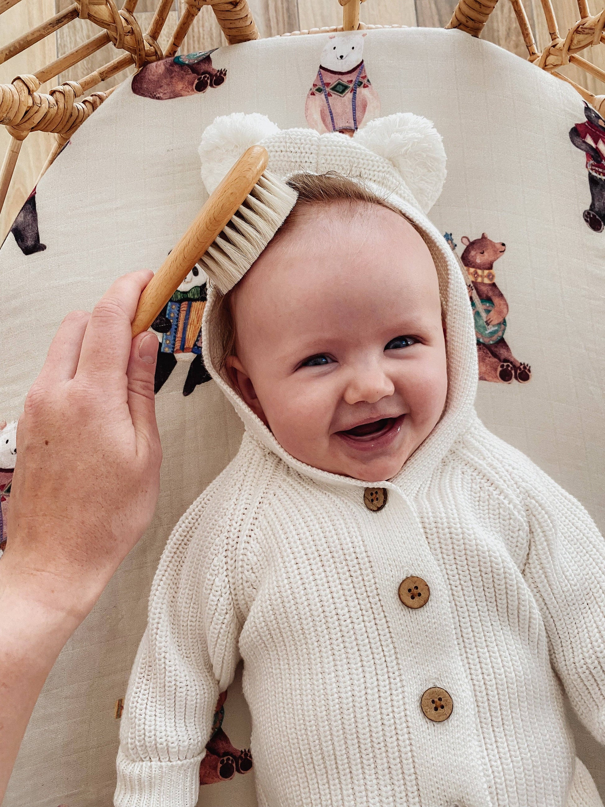 Baby in a white knit outfit with bear ears smiling at a brush held by an adult.