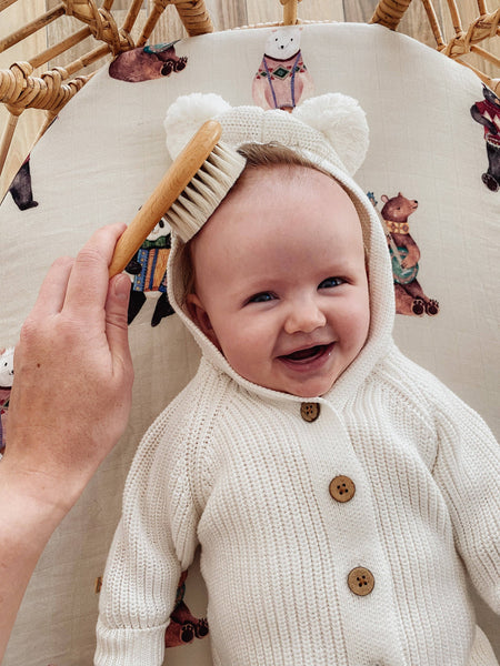 Baby in a white knit outfit with bear ears smiling at a brush held by an adult.
